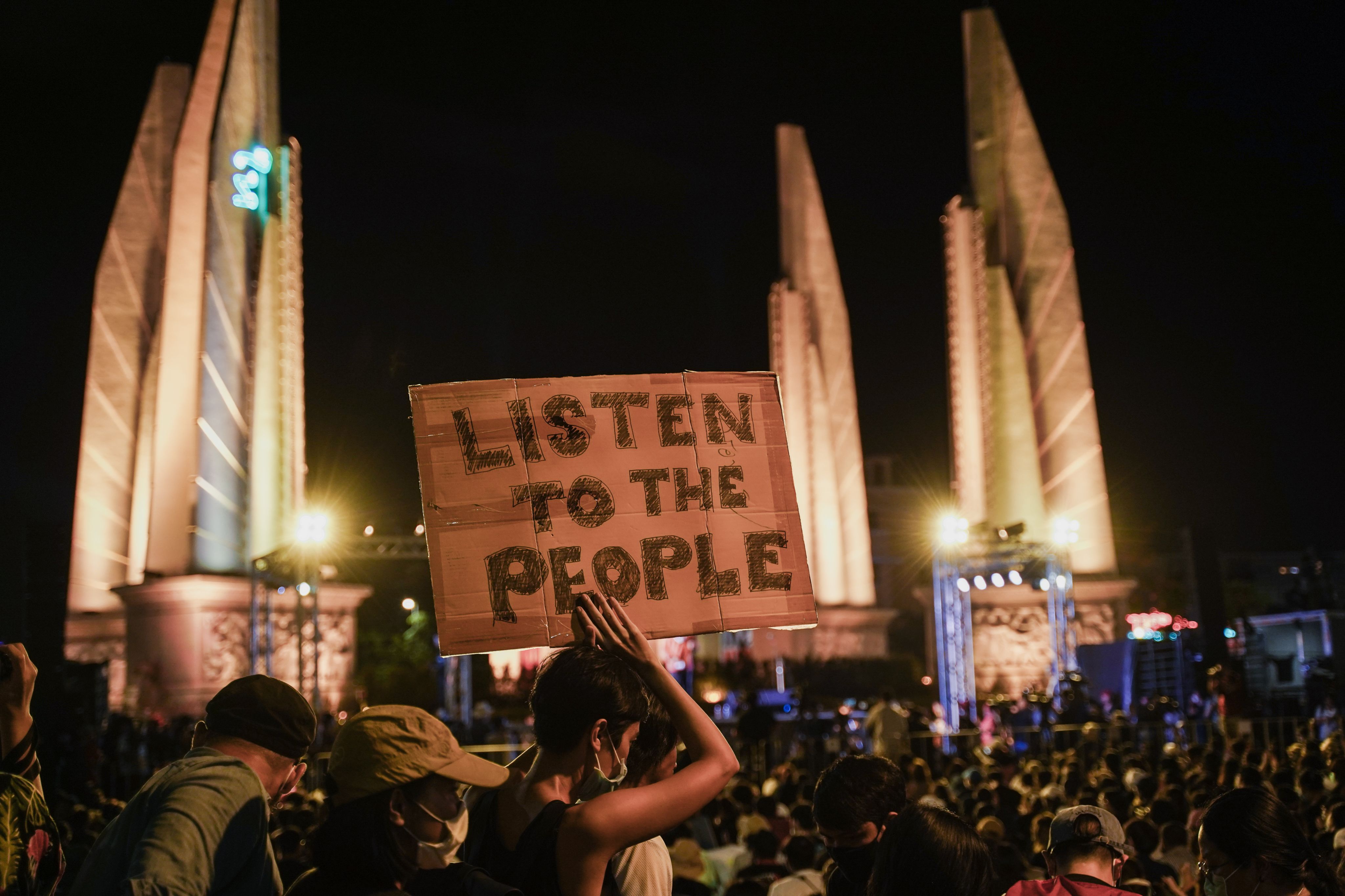 Protest in Bangkok at night time with the Democracy Monument in the background. The main focus of the photo is a woman holding a sign above her head that reads: LISTEN TO THE PEOPLE.