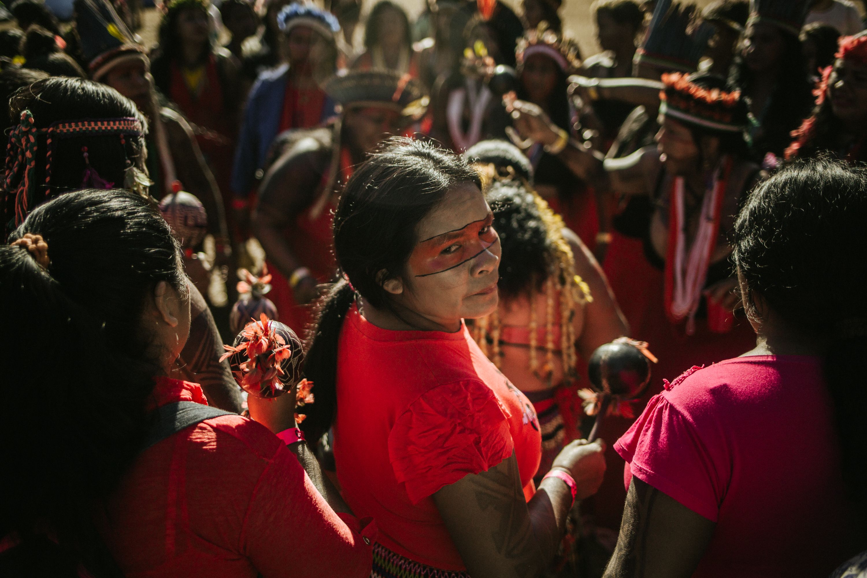 An indigenous Brazilian woman wearing red, with a red mask painted across her eyes, looks back over her shoulder at the camera. She is surrounded by other Indigenous Brazilians.